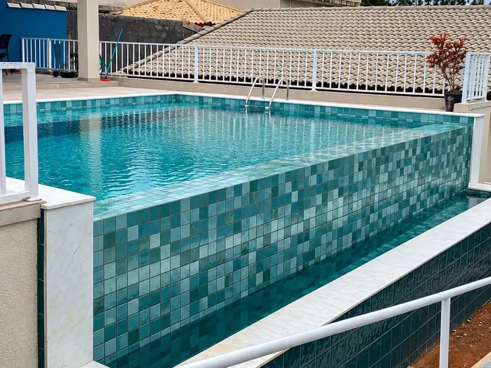 Piscina de alvenaria elevada com pastilha em tons de verde e azul por dentro e na parede externa, com guarda-corpo branco, em obra da Marks Piscinas no São Felipe, em Atibaia