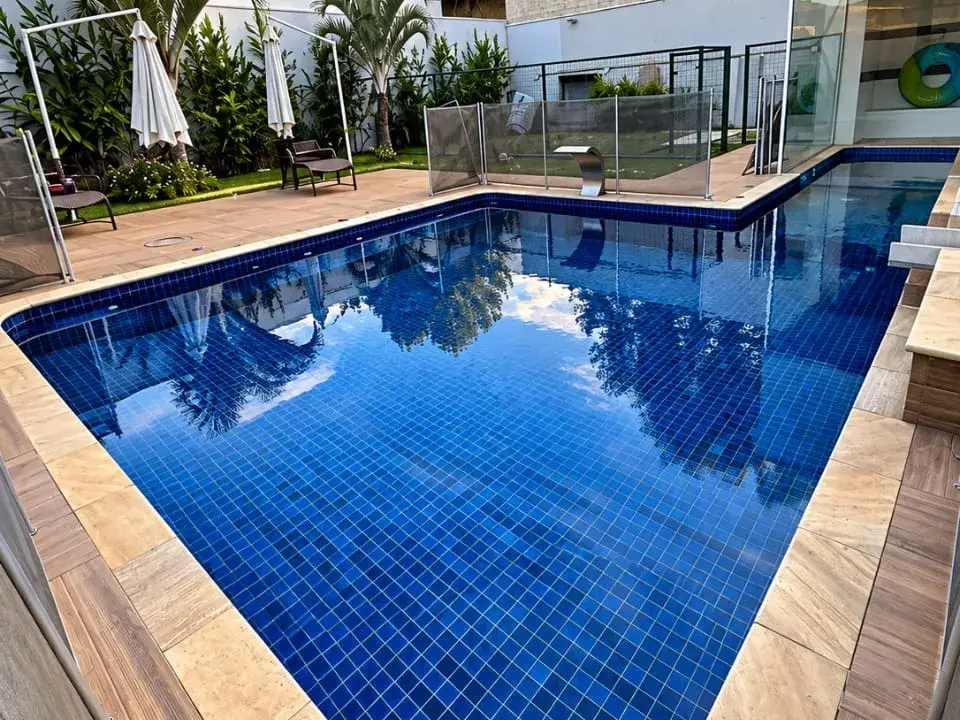 Piscina de alvenaria em formato de L com pastilha azul e borda de pedra clara