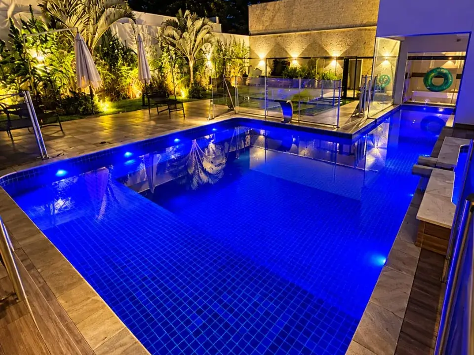 Piscina com LED azul dentro da água e jardim iluminado ao fundo, à noite